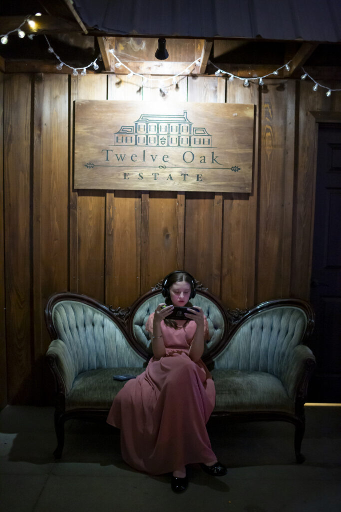 a young girl sits on a mint green vintage couch below a Twelve Oak Estate venue sign playing on her phone with headphones on